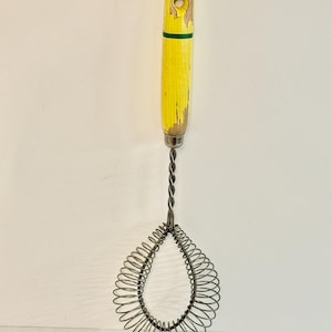 May include: Vintage egg whisk with a yellow painted wooden handle and a green stripe. The whisk's wire coils are a teardrop shape. The handle shows signs of wear, adding to its antique charm. A classic kitchen tool.