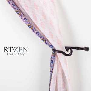 May include: Black metal curtain tieback with a decorative swirl design. The tieback is holding a pink and white patterned curtain with a blue and yellow border.
