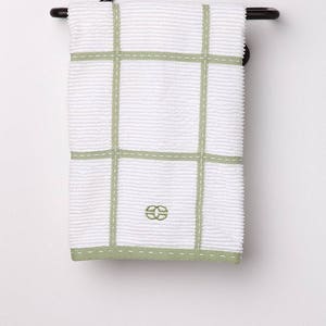 May include: A white kitchen towel with green checkered pattern and a green embroidered logo hangs on a black metal towel rack with a decorative scroll.