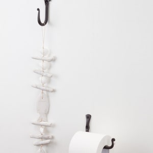 May include: Black metal toilet roll holder with a white roll of toilet paper. A black metal hook with a white ceramic fish and driftwood decoration hangs above the toilet roll holder.