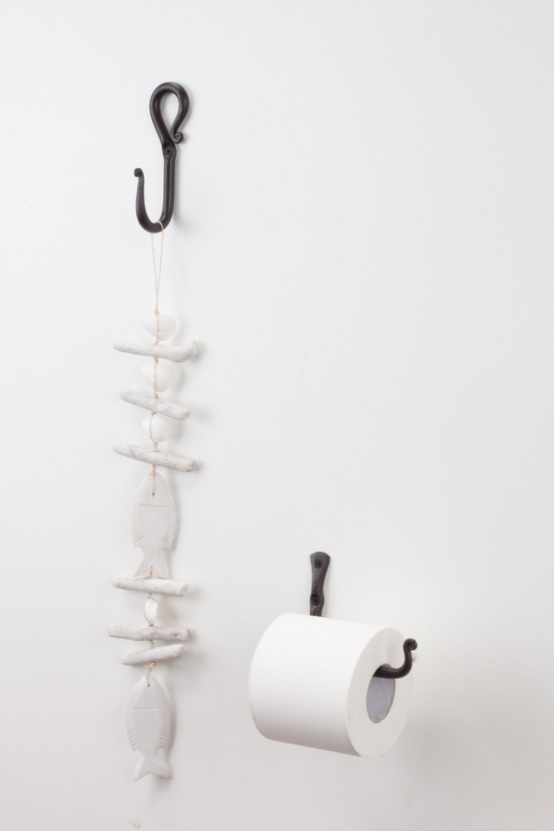 May include: Black metal toilet roll holder with a white roll of toilet paper. A black metal hook with a white ceramic fish and driftwood decoration hangs above the toilet roll holder.