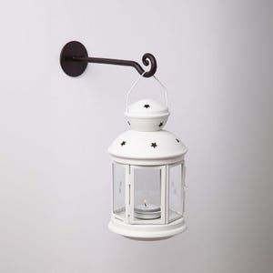 May include: A white metal lantern with a black star design hangs from a black metal hook attached to a white wall. The lantern has a glass window and a candle inside.
