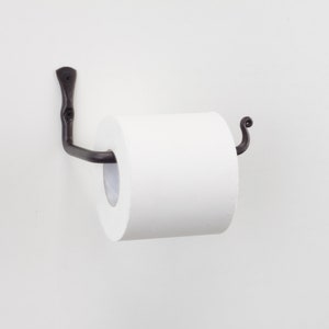 May include: Black metal toilet roll holder with a decorative scroll design. The holder is mounted to a white wall and has a roll of white toilet paper on it.