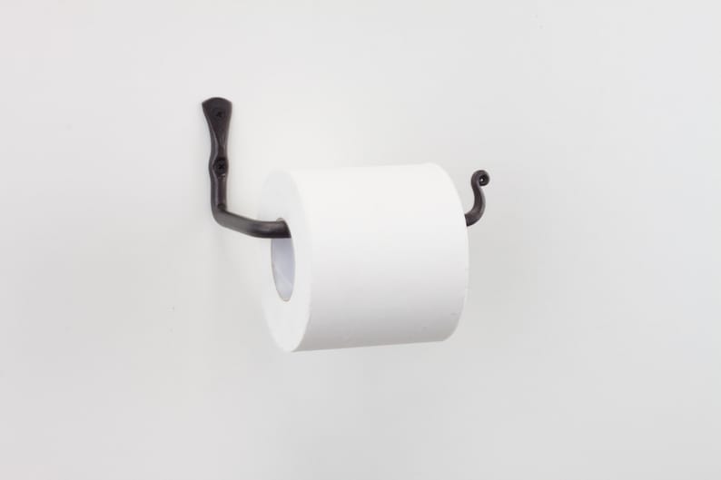 May include: Black metal toilet roll holder with a decorative scroll design. The holder is mounted to a white wall and has a roll of white toilet paper on it.