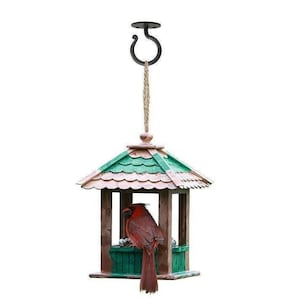 May include: A wooden bird feeder with a green roof and a red cardinal perched on the edge. The feeder is hanging from a black metal hook and a rope.