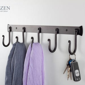 May include: A black metal wall-mounted coat rack with five hooks. The hooks are shaped like a 'J' and are curved at the top. Two scarves are hanging on the hooks. The rack is labeled "RT:ZEN Iron-Craft Decor".