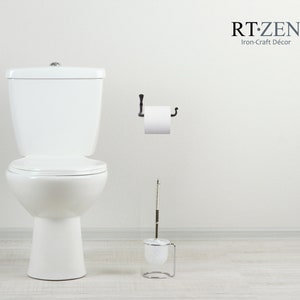 May include: White toilet with a white toilet seat and a white toilet brush in a silver holder. A roll of toilet paper is on a black metal toilet paper holder on a white wall. The text "RT.ZEN Iron-Craft D&eacute;cor" is on the wall.