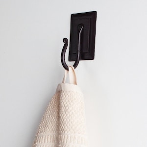 May include: A black metal hook with a decorative scroll design is mounted on a white wall. A white towel is hanging from the hook.