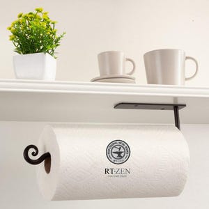 May include: Black metal paper towel holder mounted under a white shelf. The holder has a decorative scroll design and a white paper towel roll is attached. The text "RT-ZEN Iron-Craft Decor" is visible on the paper towel roll.
