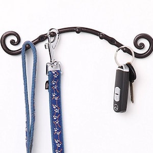 May include: A brown metal wall hook with a curved design and two decorative swirls at each end. A blue leash with a paw print pattern and a set of keys with a black key fob are hanging from the hook.