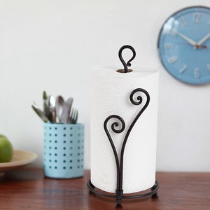 May include: A black metal paper towel holder with a decorative scroll design. The holder is holding a white paper towel roll.