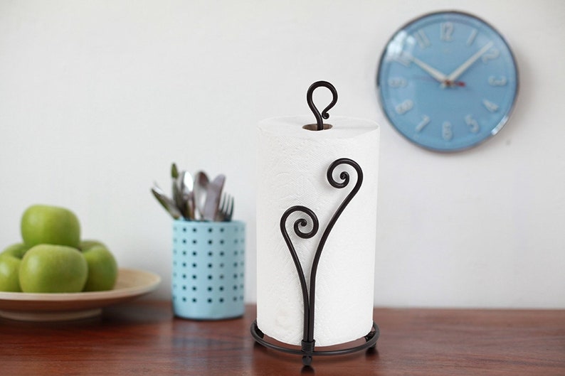 May include: A black metal paper towel holder with a decorative scroll design. The holder is holding a white paper towel roll.