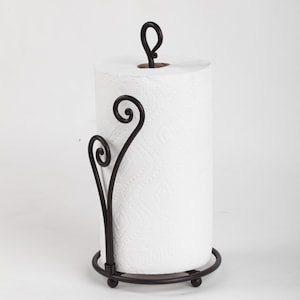 May include: Black metal paper towel holder with a decorative scroll design. The holder has a round base and a hook at the top to hold the paper towel roll.