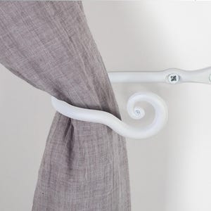 May include: White metal curtain holdback with a decorative scroll design. The holdback is attached to a wall with screws.