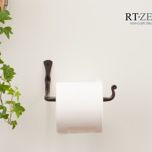 May include: A black wrought iron toilet roll holder with a scroll design on the end. The holder is mounted to a white wall and has a roll of white toilet paper on it.