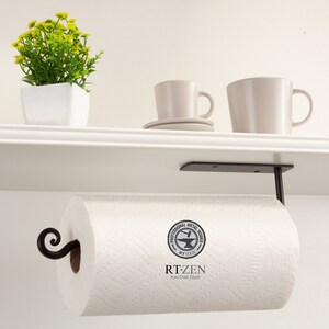Pode incluir: Suporte de papel toalha de metal preto montado sob uma prateleira branca. O suporte tem um design de rolagem decorativo e um logotipo redondo que diz "Professional Metal Works RT-ZEN Iron-Craft Decor".