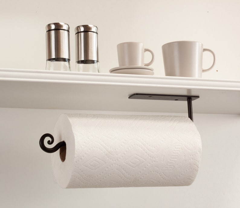 Pode incluir: Suporte de papel toalha de metal preto montado sob uma prateleira branca. O suporte tem um design de rolagem decorativo e segura um rolo de papel toalha branco.