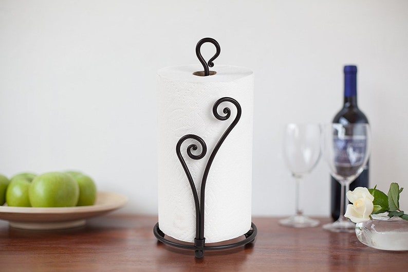 May include: Black metal paper towel holder with a decorative scroll design. The holder has a round base and a hook on top to hold the paper towel roll.