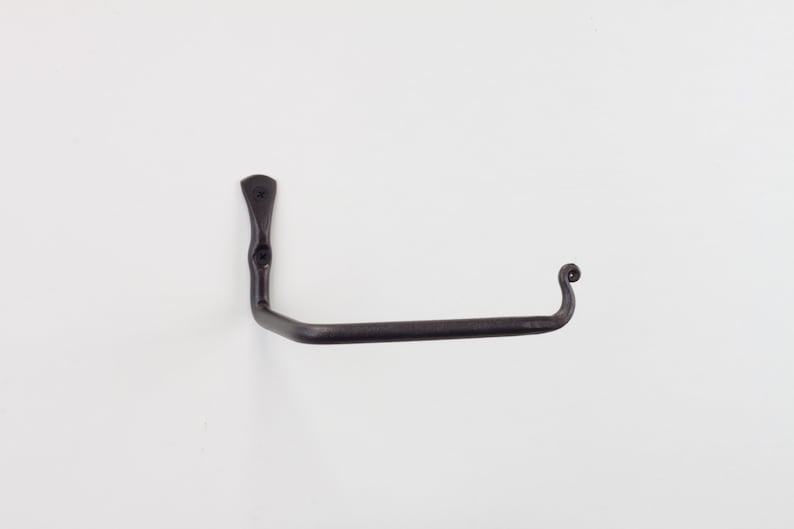 May include: A black metal toilet roll holder with a decorative scroll design on the end. The holder is mounted to a white wall.