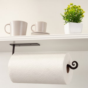 Pode incluir: Suporte de papel toalha de metal preto montado sob uma prateleira branca. O suporte tem um design decorativo de rolagem e segura um rolo de toalhas de papel brancas.