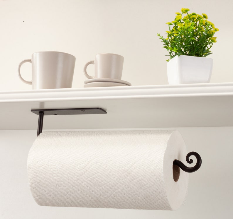 Pode incluir: Suporte de papel toalha de metal preto montado sob uma prateleira branca. O suporte tem um design decorativo de rolagem e segura um rolo de toalhas de papel brancas.