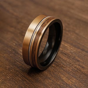 May include: A close-up of a gold-colored ring with a wood inlay and a thin silver band. The ring has a black interior and is set against a wooden surface. The ring's design features a combination of textures and colors.