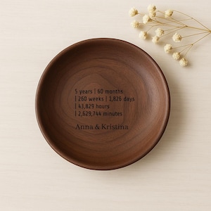 Może przedstawiać: Okrągła, ciemnobrązowa drewniana miska z wygrawerowanym tekstem upamiętniającym 5-lecie. Tekst zawiera pomiary czasu w latach, miesiącach, tygodniach, dniach, godzinach i minutach, wraz z imionami "Anna & Kristina."