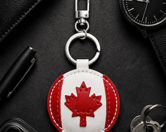 May include: A round keychain featuring the Canadian flag. The keychain has a white center with a red maple leaf, surrounded by a red and white border. It is attached to a silver keyring and clasp. Other items in the image include a watch, keys, and a phone.