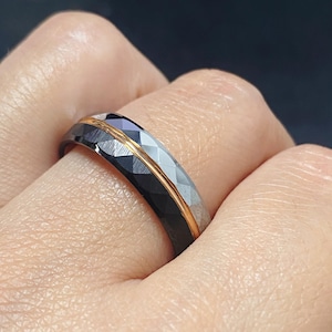 May include: A black and silver ring with a faceted design and a thin gold band. The ring is held by a person's hand, showcasing its intricate details and contrasting colours. The ring's design is geometric and modern.