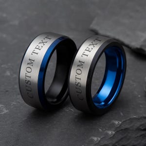 May include: Two rings with the text "CUSTOM TEXT" engraved on the outer band. One ring has a black exterior with a blue interior band, and the other has a silver exterior with a blue interior band. The rings are displayed on a dark grey surface.