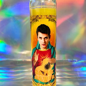 May include: A yellow candle with a picture of a man with dark hair and a red shirt holding a guitar. The text "SAINT MAYER" is printed on the candle.