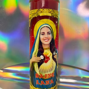 May include: Red candle with a gold trim and a picture of a woman with long dark hair. The woman is wearing a blue robe and holding a heart with a sword through it. The text "Saint Lana" is printed below the image.