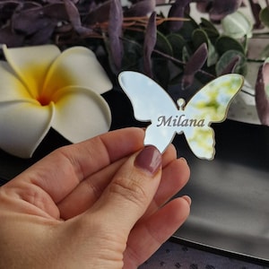 Può includere: Un segnaposto a forma di farfalla argentata con il nome "Milana" inciso. Un'ala presenta un disegno giallo e verde. Il segnaposto è tenuto in mano, con un piatto nero e fiori sullo sfondo.