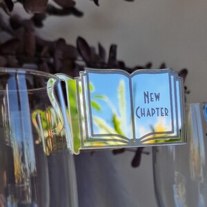 Könnte beinhalten: Klare Acryl-Getränkeanhänger in Form von offenen Büchern mit dem Schriftzug "New Chapter". Die silberfarbenen Anhänger sind an einem Sektglas befestigt. Der Hintergrund umfasst getrocknete Blumen.