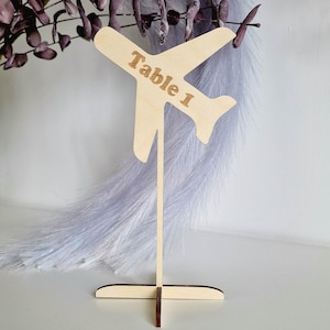 Puede incluir: Un portanúmeros de mesa de madera con forma de avión. El avión tiene grabado el texto "Table 1". El soporte es de madera clara y se apoya en una base en forma de cruz. El fondo incluye elementos decorativos morados y blancos.