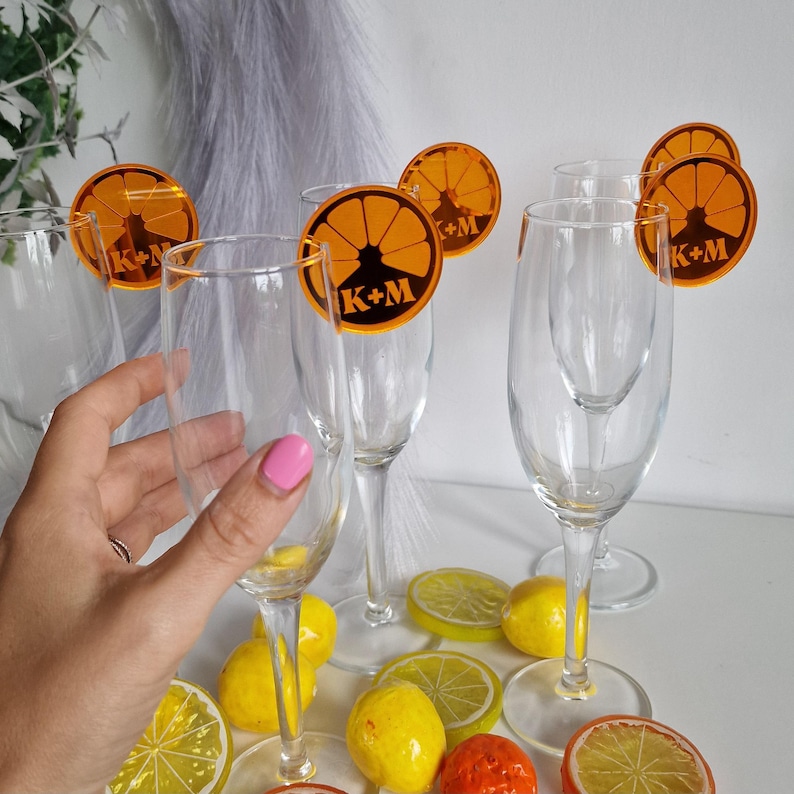 May include: Orange and gold glass charms with the text "K+M" on them. The charms are attached to champagne flutes. The flutes are sitting on a table with lemons and limes.