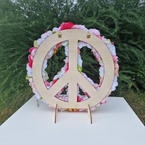 May include: A wooden peace sign with a floral wreath made of pink and white flowers. The peace sign has two holes at the top for hanging.