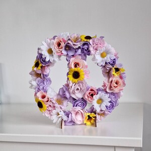 May include: A peace sign made of artificial flowers. The flowers are in shades of pink, purple, and yellow. The peace sign is on a white surface.