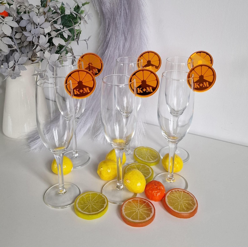 May include: Six champagne flutes with orange lemon slice place cards that say "K+M" on them. The flutes are arranged on a white surface with lemons and limes around them.