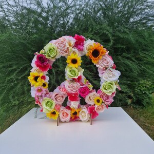 May include: A peace sign made of artificial flowers. The flowers are in shades of pink, white, yellow, and green. The peace sign is sitting on a white surface.