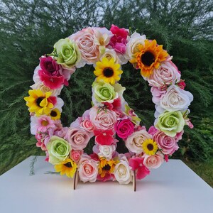 May include: A peace sign made of artificial flowers in various colors, including pink, yellow, white, and green. The flowers are arranged in a circular pattern around the peace sign.
