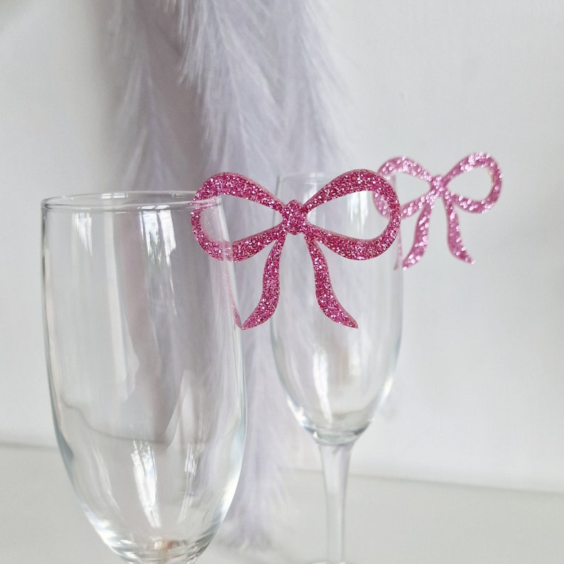 Peut inclure: Deux fl&ucirc;tes &agrave; champagne en verre transparent avec des marqueurs de boisson en forme de n&oelig;ud en paillettes roses.