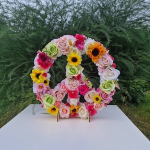 May include: A peace sign made of artificial flowers in shades of pink, yellow, white, and green. The flowers are arranged in a circle with a peace sign shape in the center.