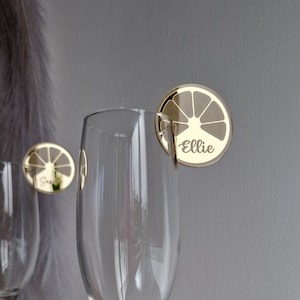 May include: Gold-colored glass markers with a lemon slice design and the name "Ellie" printed on them. The markers are attached to a clear glass.