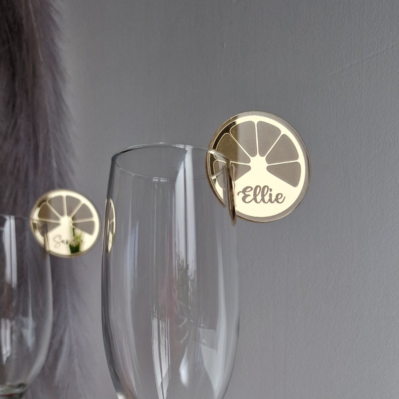 May include: Gold-colored glass markers with a lemon slice design and the name "Ellie" printed on them. The markers are attached to a clear glass.