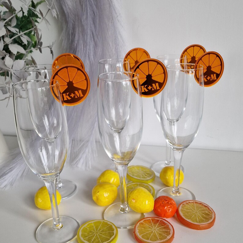 May include: Six clear glass champagne flutes with orange citrus fruit slice drink markers. Each marker has the letters "K+M" printed on it. The flutes are sitting on a white surface with yellow and orange citrus fruit slices.