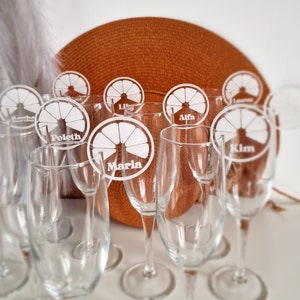May include: A set of eight clear glass champagne flutes with personalized white acrylic orange slice place cards. The place cards have the names "Poletn", "Maria", "Alfa", "Kim", "Lily", "Lauren", "Bestha", and "Laura" written on them.