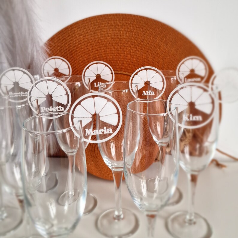 May include: A set of eight clear glass champagne flutes with personalized white acrylic orange slice place cards. The place cards have the names "Poletn", "Maria", "Alfa", "Kim", "Lily", "Lauren", "Bestha", and "Laura" written on them.