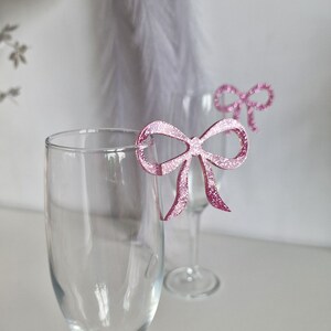 Peut inclure: Deux verres &agrave; vin en verre transparent avec des marqueurs de boisson en forme de n&oelig;ud en paillettes roses. Les n&oelig;uds sont attach&eacute;s au bord des verres.