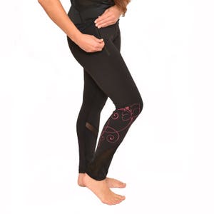 Op de afbeelding: Zwarte legging met een roze bloemenontwerp op het onderbeen. De legging heeft een mesh paneel aan de zijkant en een ritssluiting. Het model draagt een zwarte top.